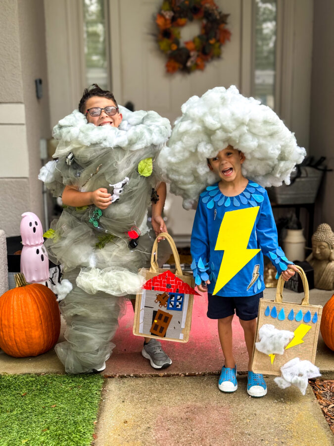Two children in creative costumes: one as a tornado with tulle, the other as a thunderstorm holding a decorated bag, with Halloween decor nearby.