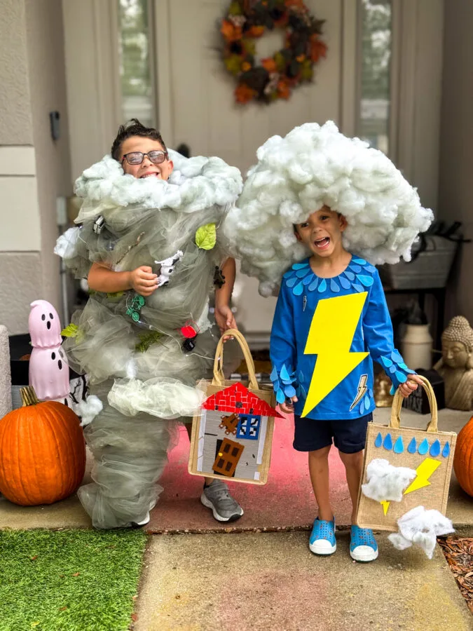 Two children in creative costumes: one as a tornado with tulle, the other as a thunderstorm holding a decorated bag, with Halloween decor nearby.