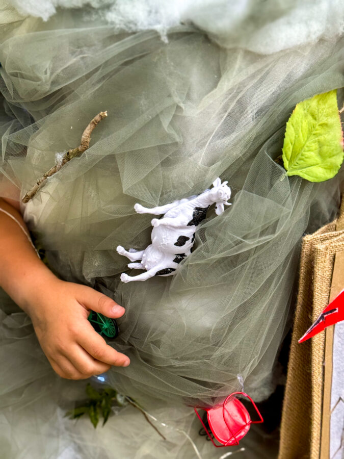 A child wearing a whimsical, cloud-themed costume adorned with faux clouds, leaves, and toy animals, standing near a pumpkin.