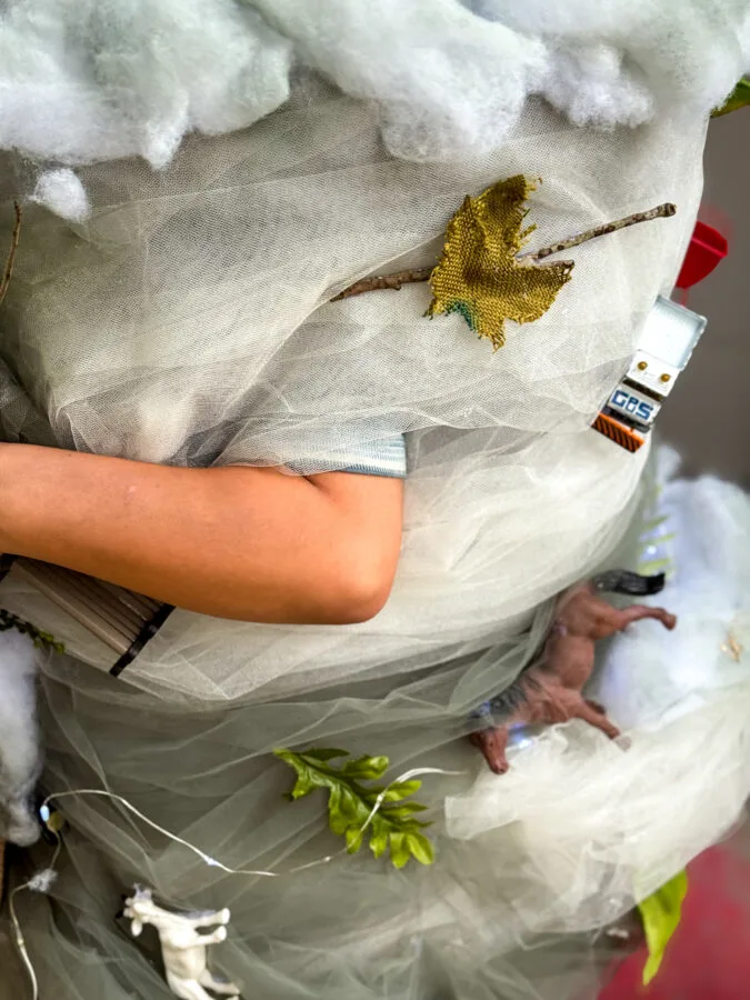 A child wearing a whimsical, tornado costume adorned with faux clouds, leaves, and toy animals, standing near a pumpkin.