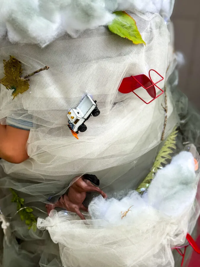 A child wearing a whimsical, tornado costume adorned with faux clouds, leaves, toy cars, and toy animals, standing near a pumpkin.
