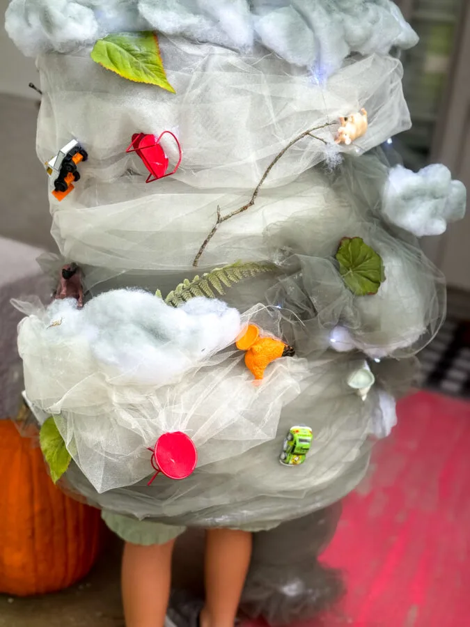 A child wearing a whimsical, tornado costume adorned with faux clouds, leaves, and toy animals, standing near a pumpkin.