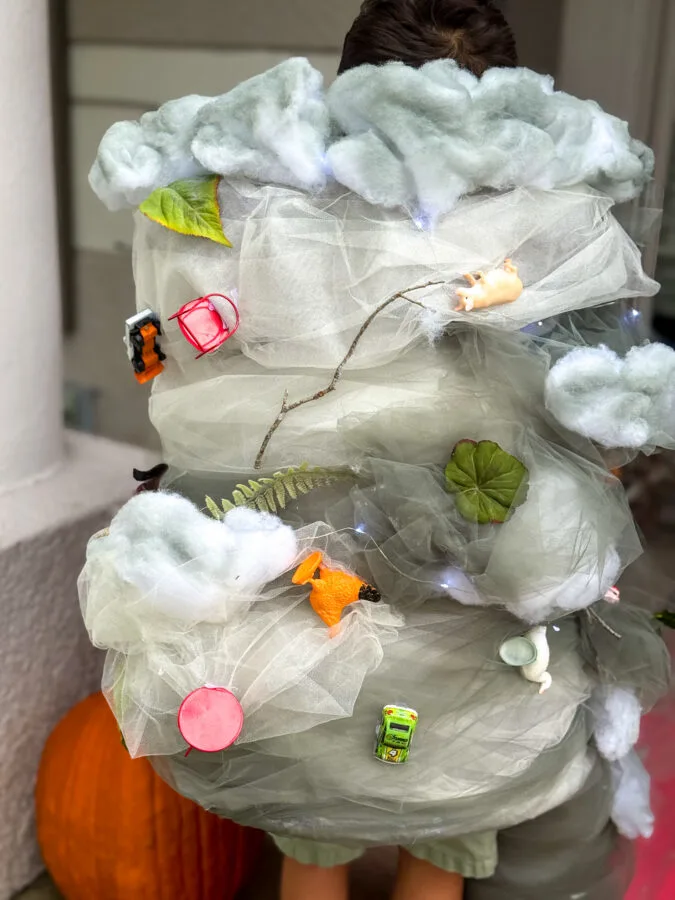 A child wearing a whimsical, tornado costume adorned with faux clouds, leaves, and toy animals, standing near a pumpkin.