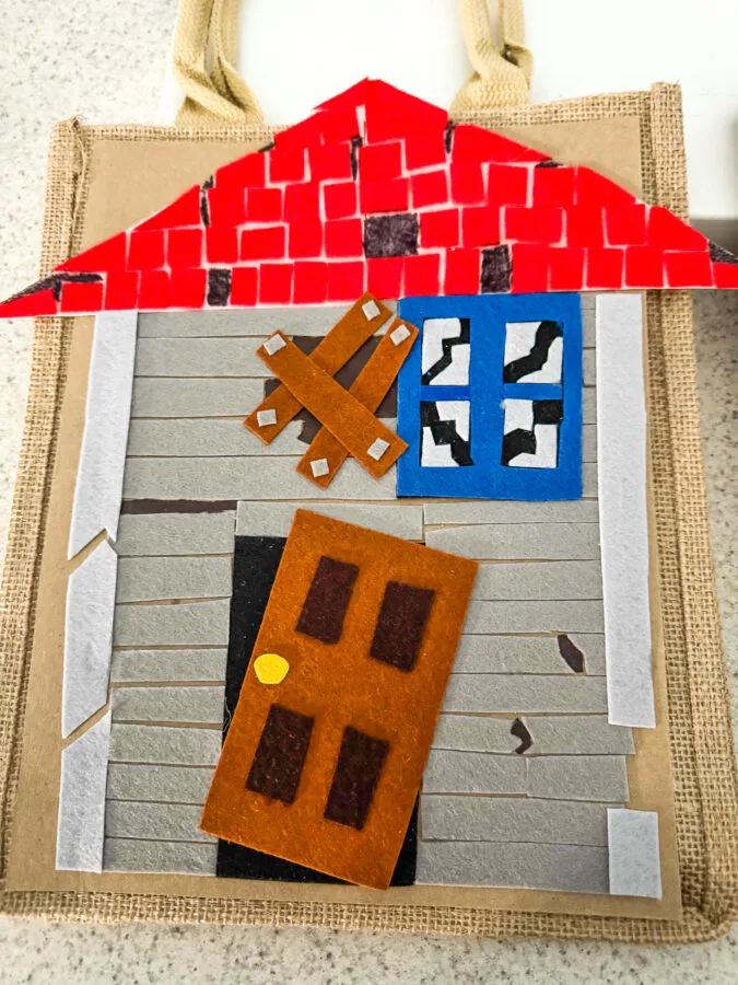 A child in a tulle costume holds a colorful felt tote bag designed like a house, featuring a red roof and various door decorations.